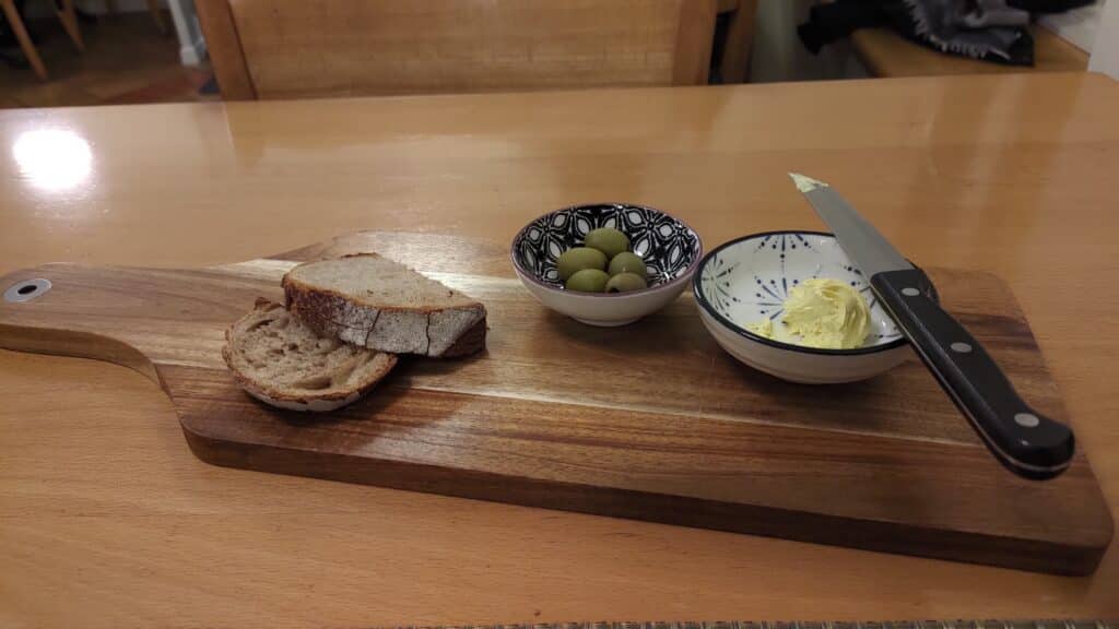 Zwei Scheiben Brot, ein Schälchen mit Oliven und eines mit Butter, hübsch auf einem Holzteller drapiert